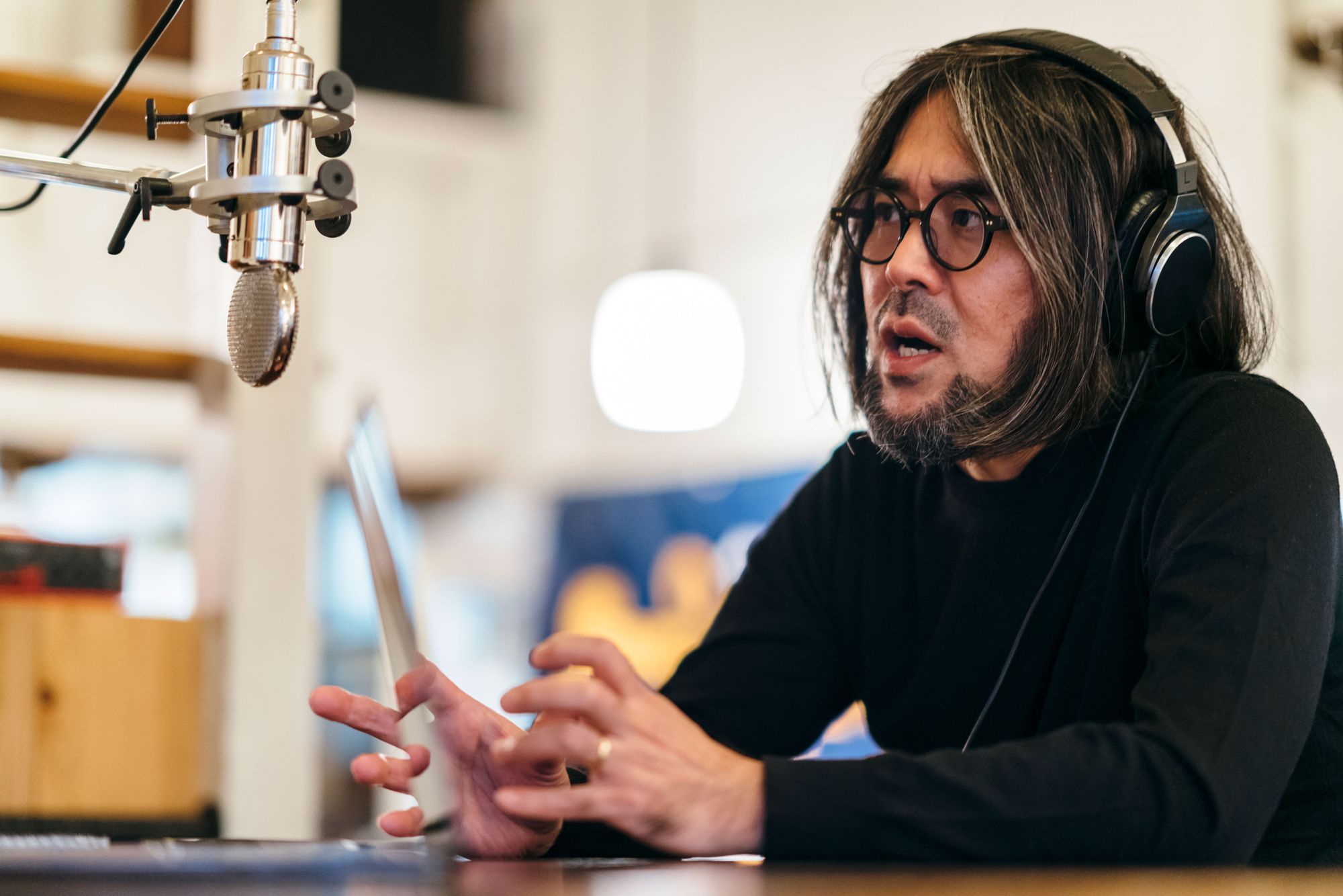 A host records in a studio, wearing headphones at a microphone with a laptop open