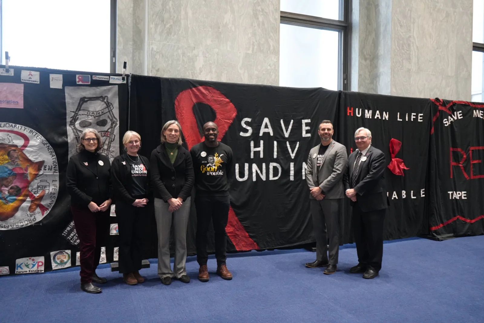 Advocates gather in front of Save HIV Funding banners inside a Capitol building hallway