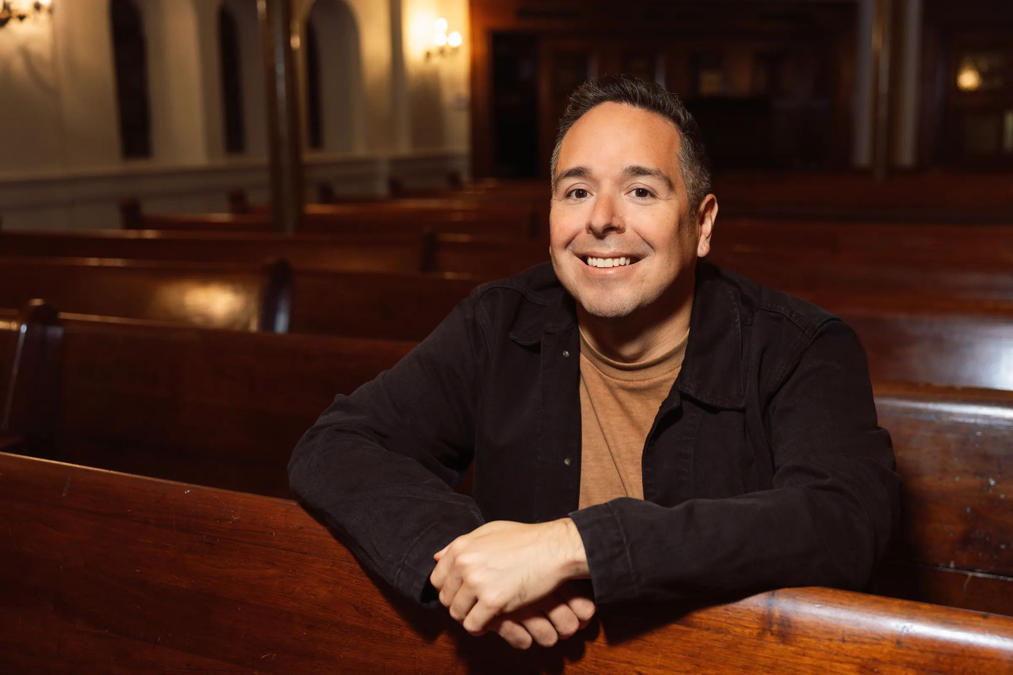 A portrait of a man smiling, seated in a wooden theater pew